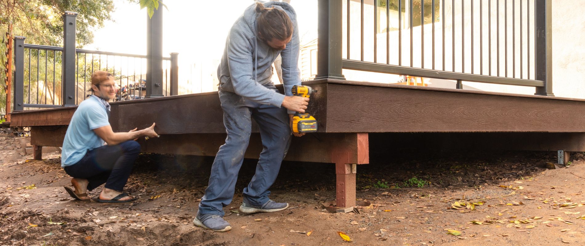 Two adults are working together to construct a wooden deck in a backyard setting. One person is using a power drill on the deck frame while the other assists nearby. The scene is shaded by a leafy tree, and the deck features black metal railings. The overall mood is industrious and collaborative, with natural daylight illuminating the workspace.