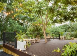 A spacious wooden deck is set outdoors beneath lush green trees, featuring a central tree growing through the platform. The area is decorated with string lights, potted plants, and contemporary outdoor furniture including chairs and a small table. The setting is tranquil and inviting, ideal for relaxation or social gatherings in a natural environment.