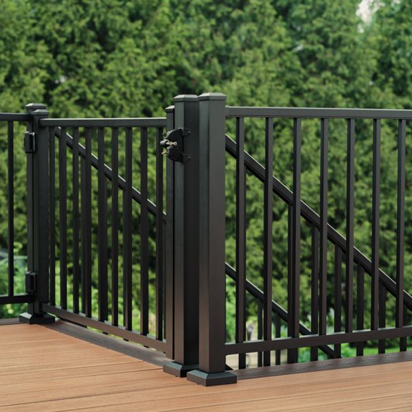 A black metal gate is installed within a matching railing on a wooden deck. The gate features vertical bars and a latch attached to a sturdy corner post. The deck surface has a warm brown tone that contrasts with the dark metal. Dense green trees fill the background, suggesting an outdoor backyard or garden setting.