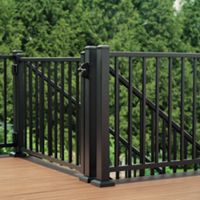 A black metal gate is installed within a matching railing on a wooden deck. The gate features vertical bars and a latch attached to a sturdy corner post. The deck surface has a warm brown tone that contrasts with the dark metal. Dense green trees fill the background, suggesting an outdoor backyard or garden setting.