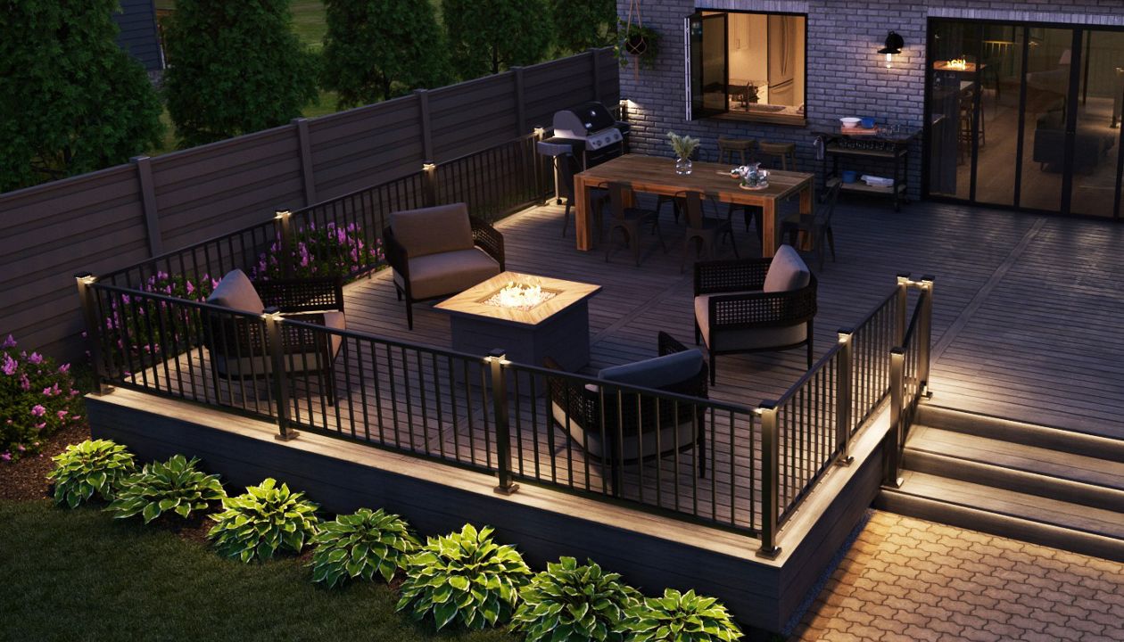 A contemporary backyard deck features a cozy seating area centered around a square gas firepit. The space is enclosed by black railings and surrounded by neatly landscaped greenery and tall evergreen trees along the fence. Warm accent lights illuminate the steps, railing posts, and the fire feature, creating an inviting atmosphere at dusk. A dining table and chairs sit near the house, visible through large sliding glass doors that open to the interior.