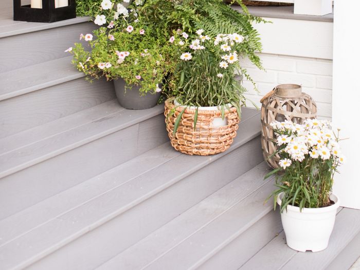 A welcoming front porch features several potted white flowers arranged on gray wooden steps. A large black lantern and a decorative candle holder add visual interest to the scene. The white railing and light siding create a bright, inviting atmosphere, while natural textures from baskets and pots enhance the cozy setting.