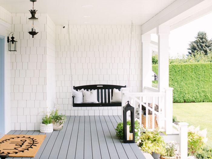 A welcoming front porch features a black wooden swing with white and patterned pillows, set against white shingle siding. The space is decorated with potted plants, a large lantern, and a cheerful doormat with visible text. The porch overlooks a lush green lawn and is bathed in natural daylight, creating a fresh and inviting atmosphere.