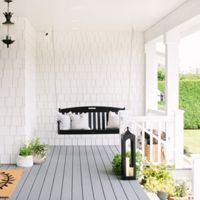 A welcoming farmhouse-style porch features a black wooden swing with white and neutral pillows. The setting includes potted plants, a large lantern, and a cheerful doormat on gray wooden flooring. The porch is surrounded by white siding and overlooks a lush green yard, creating a fresh and inviting atmosphere. Black hanging lanterns add a touch of classic charm.