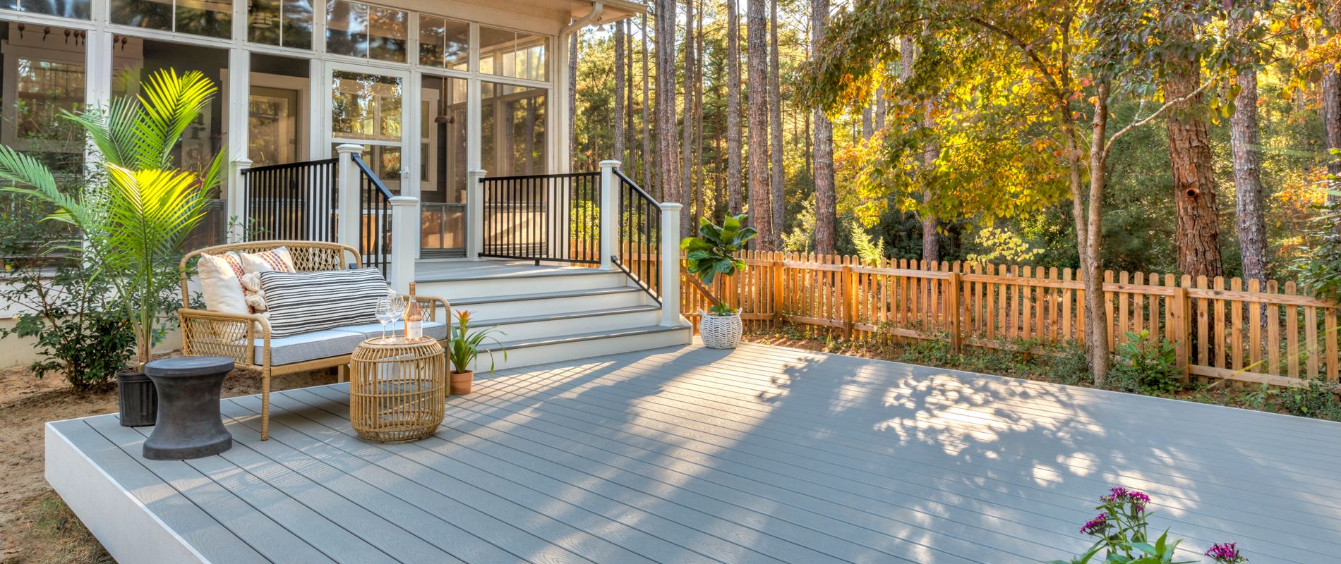 A spacious, light gray deck extends from a contemporary house, featuring comfortable wicker seating and a small table. The setting is a tranquil backyard surrounded by tall pine trees and a wooden fence, with sunlight filtering through the foliage. The area is styled for relaxation, with natural tones and cozy outdoor decor.