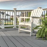 A single white Adirondack chair sits on a gray composite deck, positioned near a railing with black balusters. The setting is an outdoor residential patio or balcony, with suburban houses and green lawns visible in the background. A lush green fern adds a touch of nature, and the sky is partly cloudy, creating a calm and inviting atmosphere.