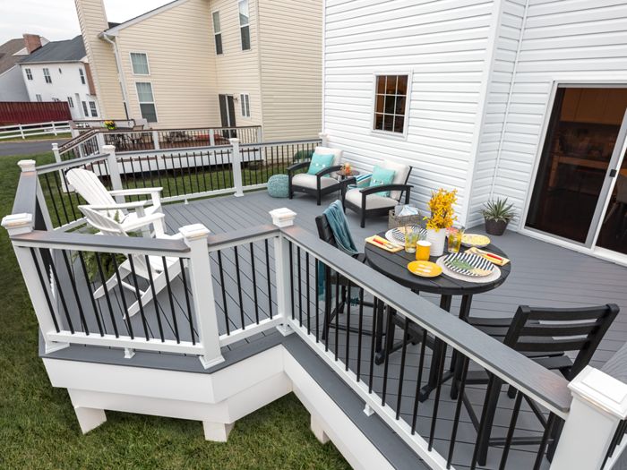 A spacious, elevated backyard deck features a dark gray floor and white railings with black balusters. The setting includes a round table set for four with plates, cups, and a yellow floral centerpiece, as well as cushioned lounge chairs and a white rocking chair. The deck is attached to a white house and overlooks a grassy yard, creating a relaxed and inviting outdoor living space.