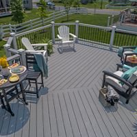 A spacious gray deck features multiple seating areas with cushioned chairs and Adirondack chairs, set up for relaxation and casual dining. Two small tables are set with plates, drinks, and snacks, while decorative plants and throws add a cozy touch. The deck overlooks a green lawn and is enclosed by white fencing, creating a welcoming outdoor living space.