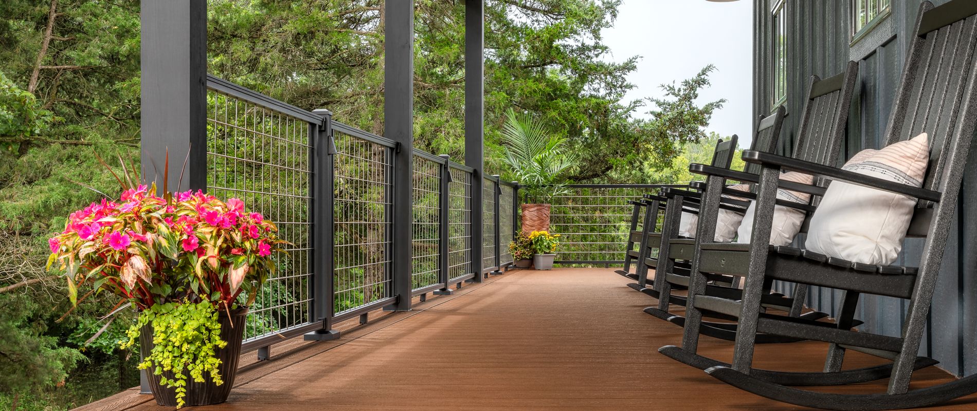 A spacious covered porch features several black wooden rocking chairs with cushions, inviting relaxation. The porch overlooks lush greenery and is decorated with vibrant potted flowers, adding a pop of color. Overhead, industrial-style lights provide illumination, and the setting exudes a peaceful, rustic charm. The scene is tranquil, perfect for enjoying nature and quiet moments.