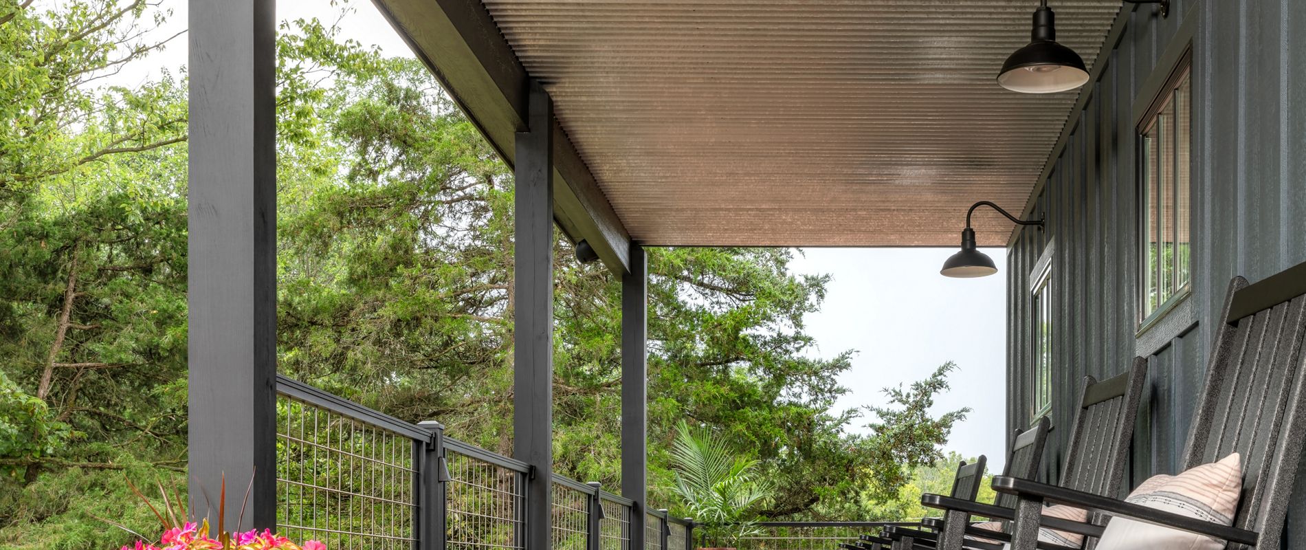 A covered porch features several black wooden rocking chairs with cushions, set against a backdrop of lush green trees. The porch has a metal roof, industrial-style lighting, and a wooden floor, creating a cozy and inviting outdoor space. Brightly colored potted flowers add a cheerful accent to the scene.