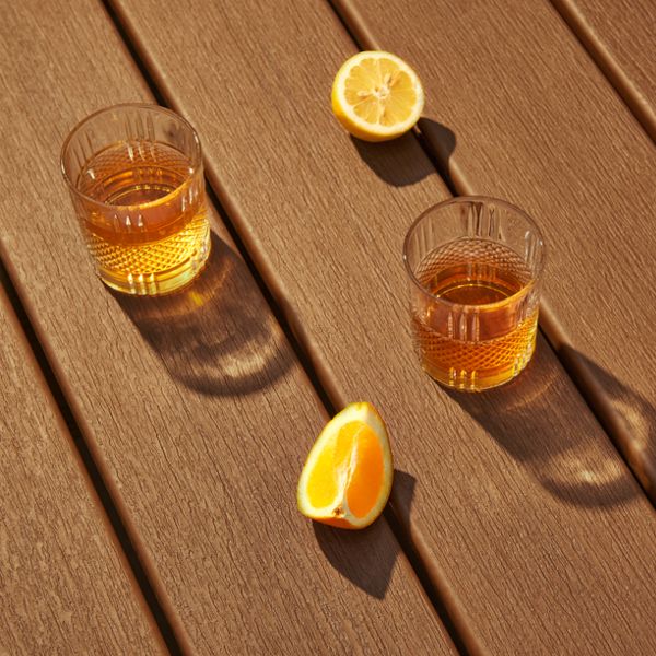 Two clear glasses filled with iced tea are placed on a wooden deck under warm sunlight. Fresh lemon slices accompany the drinks, adding a vibrant touch to the composition. The shadows cast by the glasses and lemons enhance the visual depth of the scene.