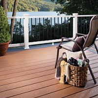 A cozy outdoor deck overlooks a serene lake surrounded by lush trees and hills. The scene features a single cushioned chair and a woven basket filled with blankets and a book, suggesting relaxation. Warm-toned composite decking contrasts with the white and black railing, while soft sunlight enhances the tranquil mood. No visible people or text are present in the image.