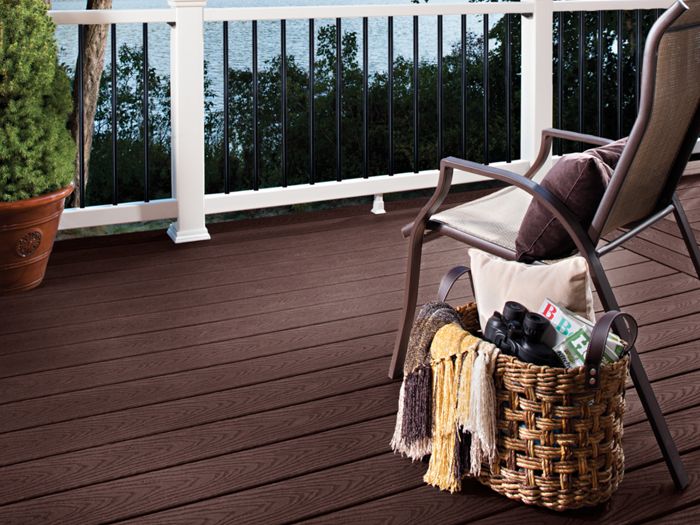 A cozy outdoor deck overlooks a tranquil lake surrounded by lush green forest and hills. The deck features dark wood planks, a white and black railing, and a comfortable chair with a blanket. A woven basket with a drink bottle, fruit, and a scarf sits nearby, creating a relaxing and inviting atmosphere.