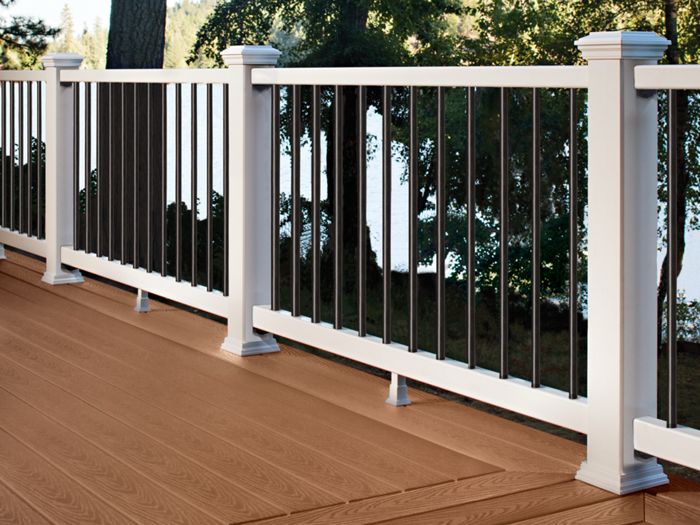 A spacious outdoor deck features wood-look composite flooring and a sturdy white railing with black balusters. The deck overlooks a lush, green forest with tall trees and a glimpse of water in the background. The setting is serene and natural, ideal for relaxation or outdoor gatherings. The deck boards have a warm brown tone and visible wood grain texture.
