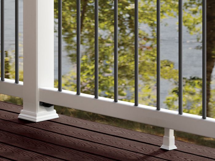 A white vertical railing with dark metal balusters is installed on a deck with rich brown wood planks. The deck overlooks a serene lake surrounded by lush green trees, creating a peaceful outdoor setting. Sunlight filters through the foliage, casting natural light and shadows on the scene. No visible text, numbers, or logos are present in the image.