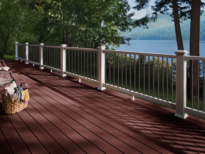A spacious wooden deck overlooks a tranquil lake surrounded by trees. The deck features rich brown planks and a white railing, with comfortable seating and a woven basket visible. Sunlight casts shadows across the deck, creating a peaceful outdoor setting ideal for relaxation.