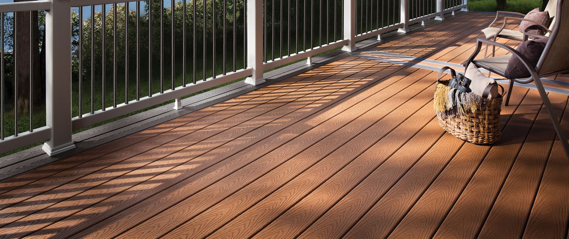 A spacious wooden deck overlooks a serene lake, bordered by a white railing. Two lounge chairs and a woven basket are positioned for relaxation, with sunlight casting shadows across the deck. The setting is surrounded by lush green trees, creating a peaceful outdoor retreat.