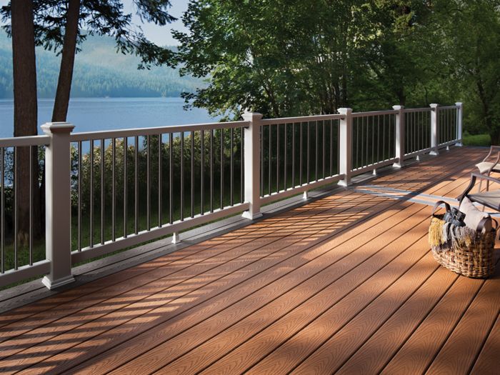 A spacious wooden deck overlooks a serene lake, surrounded by lush green trees. The deck features comfortable seating and a woven basket, creating a relaxing outdoor setting. Sunlight casts shadows through the railing, enhancing the warm, inviting atmosphere. The scene suggests leisure and tranquility in a natural environment.