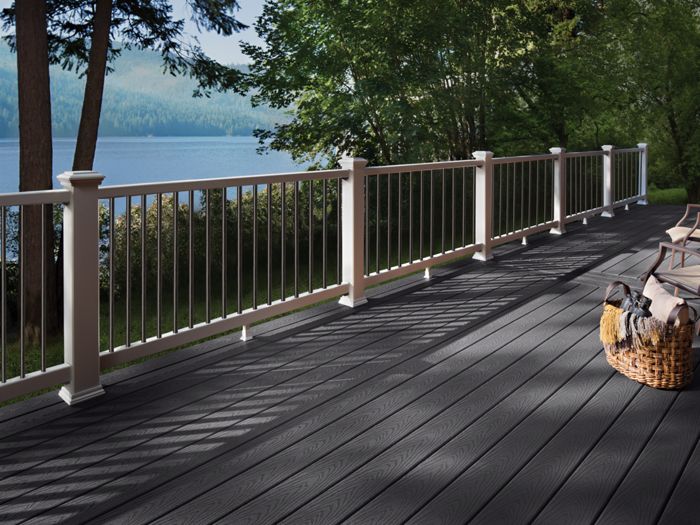 A spacious outdoor deck overlooks a serene lake surrounded by trees. The deck features dark composite flooring, white railings, and comfortable seating with cushions. A woven basket and soft shadows add to the tranquil, inviting atmosphere, perfect for relaxation or gatherings.