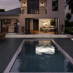 A contemporary two-story villa features a sleek rectangular swimming pool in the foreground. The open-plan living and kitchen area is visible through large glass doors, warmly lit against the dusk sky. Outdoor lounge chairs and a dining table sit on the expansive deck surrounding the pool. Palm trees and manicured greenery frame the property, creating a tranquil, upscale resort atmosphere.