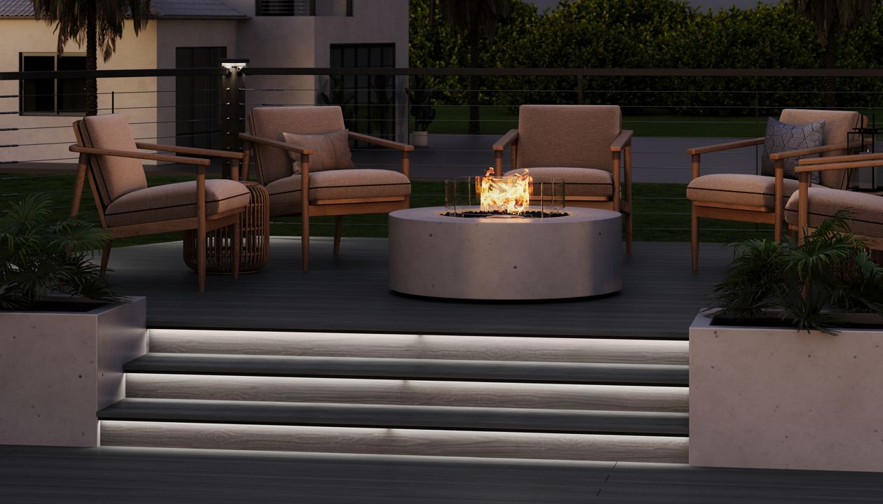 A contemporary outdoor seating area features several cushioned armchairs arranged around a circular fire pit. The scene is set on a sleek deck with illuminated steps leading up to the lounge space. A modern house and tall palm trees are visible in the background, suggesting a warm, residential setting. Soft lighting and the glow of the fire create a cozy, evening atmosphere.