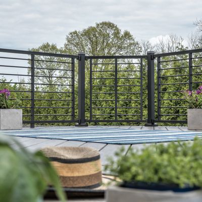 A contemporary outdoor deck features a sleek black metal railing and several potted plants. The setting is tranquil, with greenery and trees visible in the background under a cloudy sky. A striped rug and a straw hat add a relaxed, casual touch to the scene. The overall mood is peaceful and inviting, ideal for leisure or small gatherings.