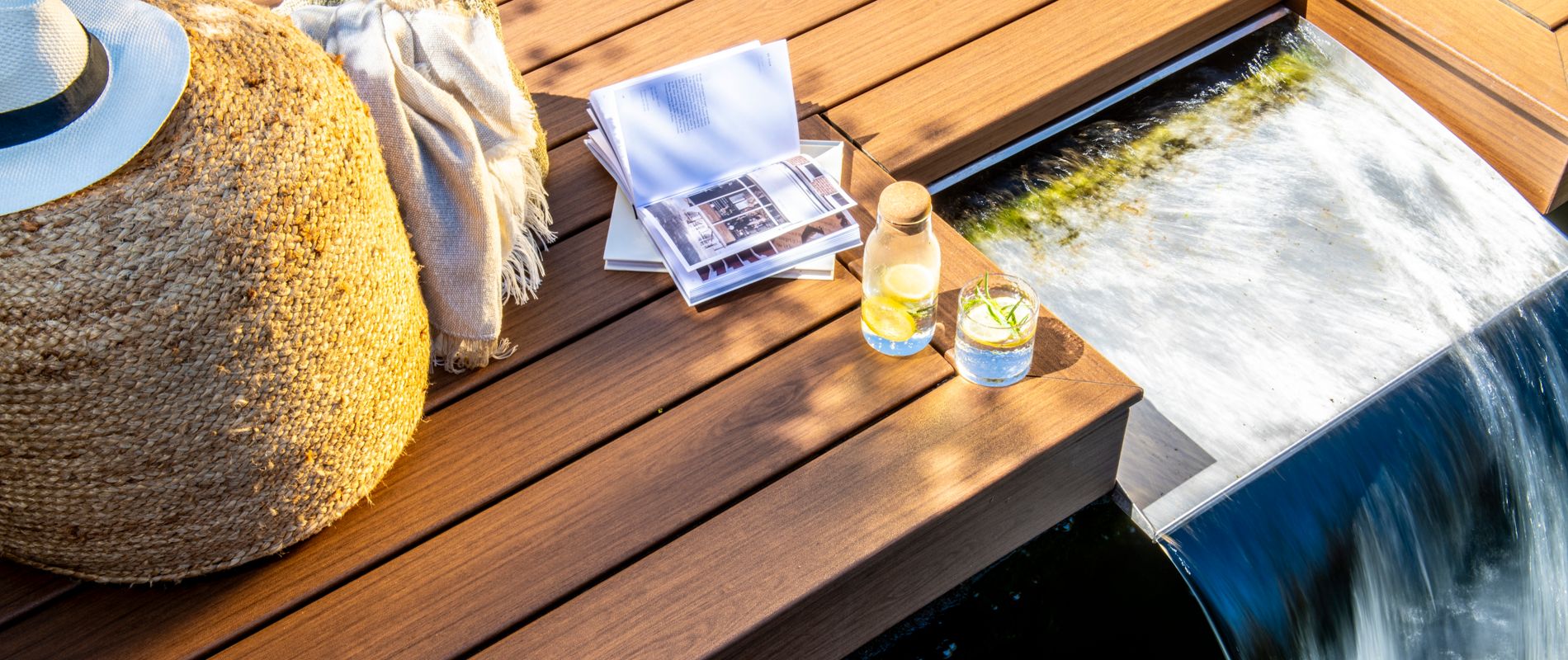 A serene outdoor deck featuring a cascading waterfall alongside wooden paneling. Decor elements include a woven basket, a sun hat, and folded linens, creating a relaxing ambiance. Two glasses with citrus-infused water and a clear bottle add a refreshing touch to the scene.