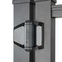 This image features a close-up view of a D&D Technologies gate hinge mounted between two vertical metal fence posts. The hinge is prominently displayed, showing its robust construction and modern design. The setting is neutral with a focus on the hardware, and the text 'D&D CLOSE' is clearly visible on the hinge. The overall palette is monochromatic with shades of gray and black.
