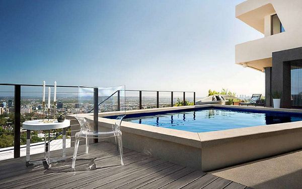 A contemporary rooftop pool is featured on a high-rise building, overlooking a sprawling cityscape under a clear blue sky. The scene includes a transparent chair and a small round table set with two white candles, creating a minimalist and elegant atmosphere. Glass railings surround the pool area, enhancing the open and airy feel. The setting exudes a calm, luxurious mood with a neutral color palette and clean architectural lines.