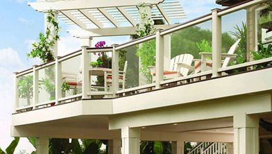 A contemporary outdoor deck features clear glass railings and white framing, creating an open and airy feel. The upper level includes a pergola with climbing plants and a seating area with white chairs and a table. Lush greenery surrounds the structure, enhancing the tranquil and inviting atmosphere.