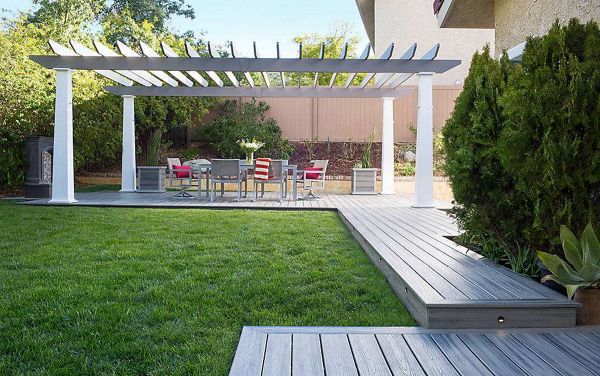A contemporary backyard features a wooden deck leading to a patio area with a white pergola. Under the pergola, there is a dining table surrounded by colorful chairs, set for outdoor gatherings. The space is bordered by lush green grass, manicured shrubs, and a privacy fence, creating a welcoming and relaxed atmosphere.