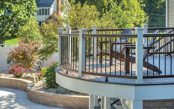 A contemporary outdoor deck features a curved metal railing and several lounge chairs, overlooking a landscaped garden. The setting is a residential backyard with lush greenery and flowering shrubs. Sunlight highlights the clean lines and inviting atmosphere, creating a relaxing space for outdoor leisure. No visible text or numbers are present in the image.