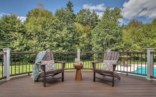 Two wooden Adirondack chairs with patterned cushions are arranged on a spacious deck, separated by a small round table. The deck overlooks a lush green backyard with a swimming pool visible in the background. A light blanket is draped over one chair, and the scene is set under a bright blue sky with scattered clouds, creating a relaxed and inviting outdoor atmosphere.