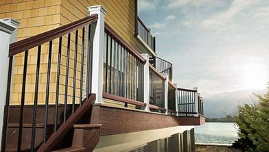 A contemporary deck with dark metal and wood railings is attached to a house overlooking a body of water. The setting features a serene waterfront view with sunlight breaking through clouds. The house exterior is clad in tan siding, and the deck is elevated with stairs leading down to a gravel area. No visible text or numbers are present in the image.