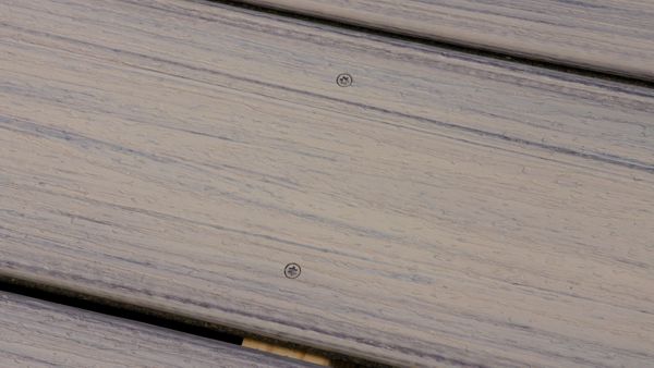 This image shows a close-up view of composite wood decking boards. The surface features a subtle wood grain texture and two visible screws securing the planks. The color palette is a blend of light gray and beige tones, giving a modern and clean appearance. No visible text or numbers are present in the image.
