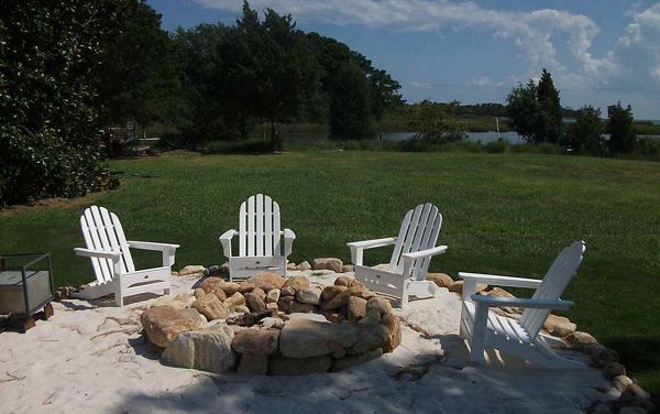 Four white Adirondack chairs are arranged in a circle around a stone fire pit on a sandy patio. The setting is an open grassy backyard with trees and a pond visible in the background. The scene is bright and sunny, evoking a relaxed and inviting outdoor atmosphere. No visible text or numbers are present in the image.