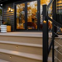 A contemporary outdoor deck features a wooden sofa with plush cushions and two sculptural white side tables. The setting is illuminated by small step lights and warm reflections from large glass doors, creating a welcoming atmosphere. Trees and greenery are visible in the background, enhancing the tranquil, modern vibe. The scene is captured at sunset, highlighting the inviting and stylish design.