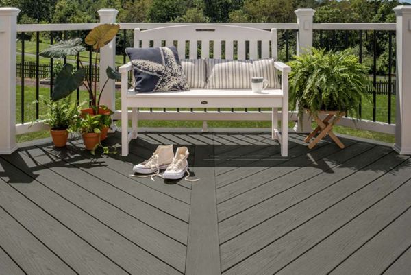 A white bench with striped cushions sits on a dark gray composite deck, surrounded by potted plants and a pair of sneakers. The setting is an outdoor patio or backyard, with a lush green landscape visible beyond the railing. The scene conveys a relaxed, inviting mood with natural light and soft, neutral tones.