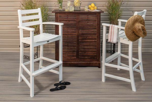Two white bar chairs are arranged in front of a wooden outdoor bar cabinet. The setting includes a summery atmosphere with lemonade, glasses, and lemons on the bar, as well as potted plants in the background. A straw hat and striped towel hang on one chair, while black flip-flops are placed on the deck floor. The overall mood is relaxed and inviting, ideal for outdoor gatherings.