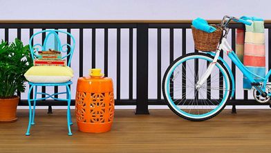 A vibrant outdoor patio features a turquoise metal chair with a yellow cushion and stacked books, next to an orange decorative side table holding a yellow teacup. A white bicycle with a basket and multicolored blanket leans against a black railing. The setting is bright and cheerful, with a potted green plant adding a fresh touch.