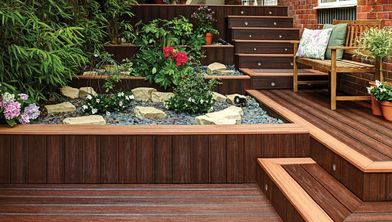 A multi-level wooden deck features integrated steps and a landscaped rock garden with lush green plants and colorful flowers. The setting is an outdoor patio adjacent to a brick house, with a wooden bench adorned with patterned cushions and potted plants. The overall mood is tranquil and inviting, with natural tones and a touch of cozy decor.