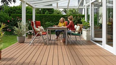 Two women are seated at a table on a spacious wooden patio deck under a clear roof. The setting is a bright, well-maintained backyard garden with lush greenery and comfortable outdoor furniture. The mood is relaxed and casual, with natural light enhancing the warm tones of the deck and chairs. No visible text or numbers are present in the image.