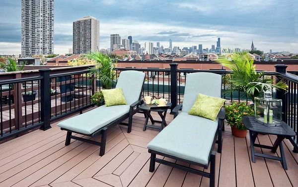 Two cushioned lounge chairs with green pillows are arranged on a wooden rooftop deck overlooking a city skyline. The setting includes potted plants, a small table with drinks, and a lantern, creating a relaxing outdoor atmosphere. Tall buildings and a dramatic sky provide an urban backdrop, while the deck's railing frames the scene. No visible text or numbers are present in the image.