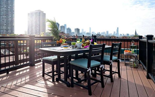 A modern outdoor dining table with four cushioned chairs is arranged on a spacious rooftop deck. The setting overlooks a vibrant city skyline under a bright, clear sky. The table is set with food, drinks, and decorative items, creating a welcoming atmosphere for gatherings. The deck features wood-style flooring and black railings, with greenery and fruit adding pops of color.
