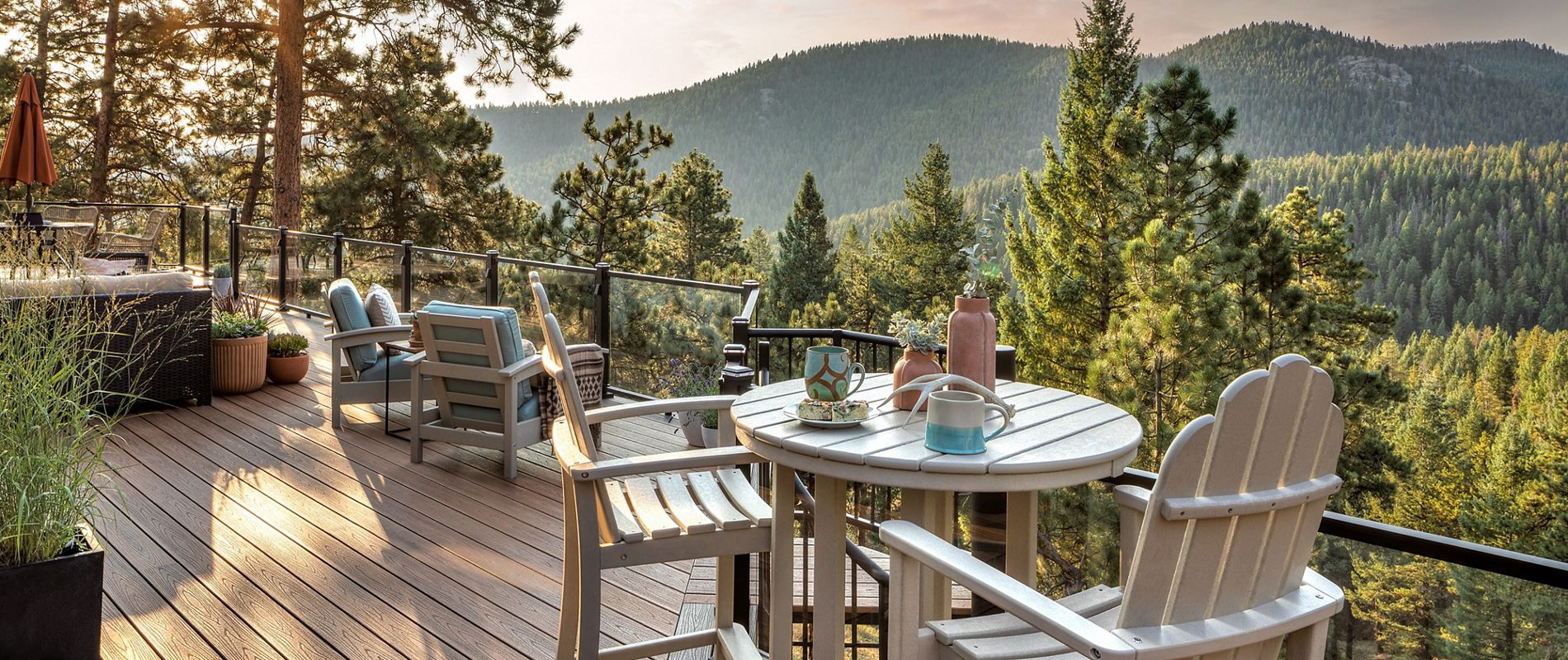A spacious wooden deck overlooks a scenic mountain landscape, surrounded by lush pine trees. The setting features several Adirondack chairs and tables, with cups and a thermos placed on one table, suggesting a relaxed outdoor gathering. The warm sunlight and soft shadows create a tranquil, inviting atmosphere, perfect for enjoying nature and quiet moments.