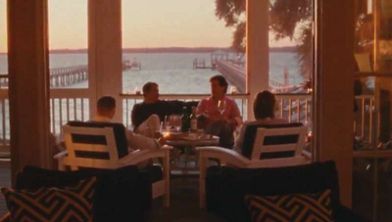 Four adults are seated on a porch, enjoying a relaxed conversation as the sun sets over a scenic waterfront. The setting features comfortable white chairs, patterned pillows, and a table with drinks, creating a cozy and inviting atmosphere. Large windows frame the view of the water, dock, and silhouetted trees, enhancing the tranquil mood. The warm, golden light of sunset fills the space, emphasizing a peaceful and social ambiance.