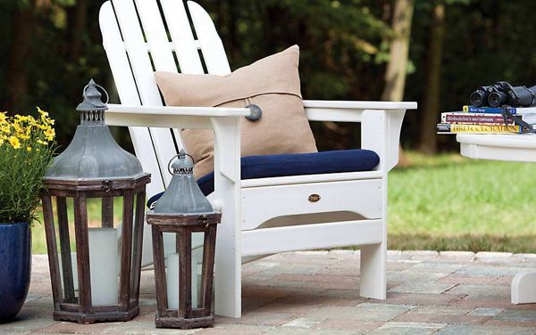 A white Adirondack chair with a navy cushion and beige pillow sits on a brick patio. Two large metal and glass lanterns are placed nearby, along with a blue pot of yellow flowers. The setting is outdoors, with a green lawn and trees in the background, evoking a relaxed and inviting atmosphere.