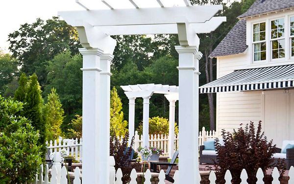 A white pergola stands prominently in a landscaped backyard, surrounded by a white picket fence and lush greenery. The scene includes outdoor seating and a house with a striped awning, creating a welcoming and relaxed atmosphere. The image features bright daylight and a clean, classic architectural style.