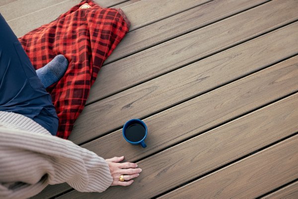 Person sitting on a checkered red and black blanket on a deck made of Trex Transcend Lineage® in the color Biscayne with their hand resting on the deck next to a mug of coffee. 