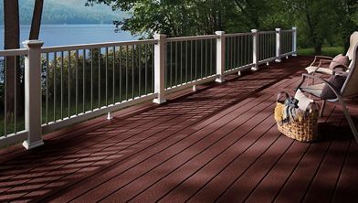 A large wooden deck overlooks a serene lake, surrounded by lush green trees. The deck features white railings and comfortable seating, including chairs and a woven basket. Sunlight casts shadows across the reddish-brown deck boards, creating a peaceful and inviting atmosphere. No visible text or numbers are present in the image.